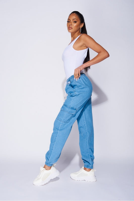 Model wears a loose fit light blue patch pocket cuffed denim joggers. Model wears a white bodysuit and white trainers.  Model stands to the side but turns her head to look into the camera. 