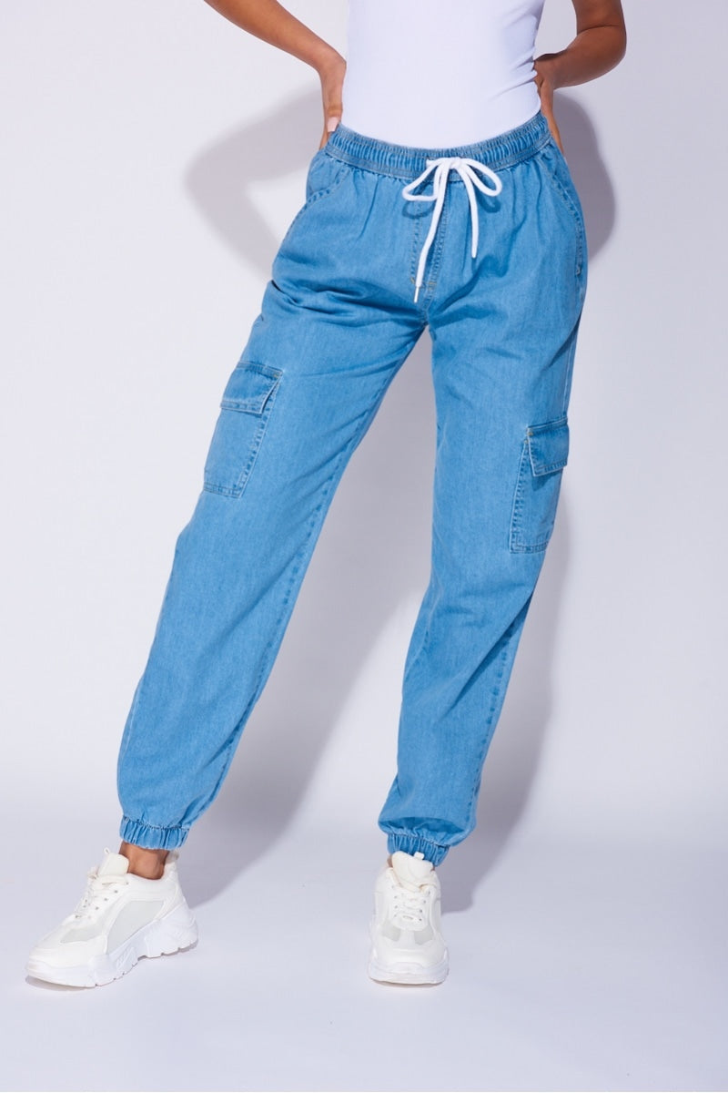 Model wears a loose fit light blue patch pocket cuffed denim joggers. Model wears a white bodysuit and white trainers.  The photograph shows a closeup of the joggers. 