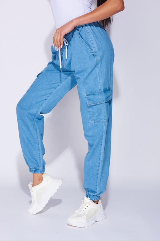 Model wears a loose fit light blue patch pocket cuffed denim joggers. Model wears a white bodysuit and white trainers.  Photograph sows a closeup of the trousers with the model standing to the side and one leg raised. 