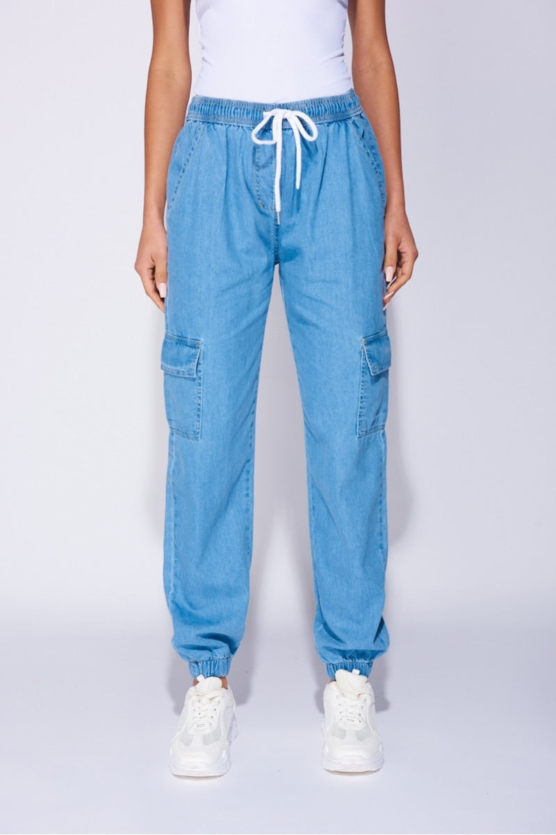 Model wears a loose fit light blue patch pocket cuffed denim joggers. Model wears a white bodysuit and white trainers.  Photograph shows a closeup of tghe trousers with the model with her hands behind her side. 