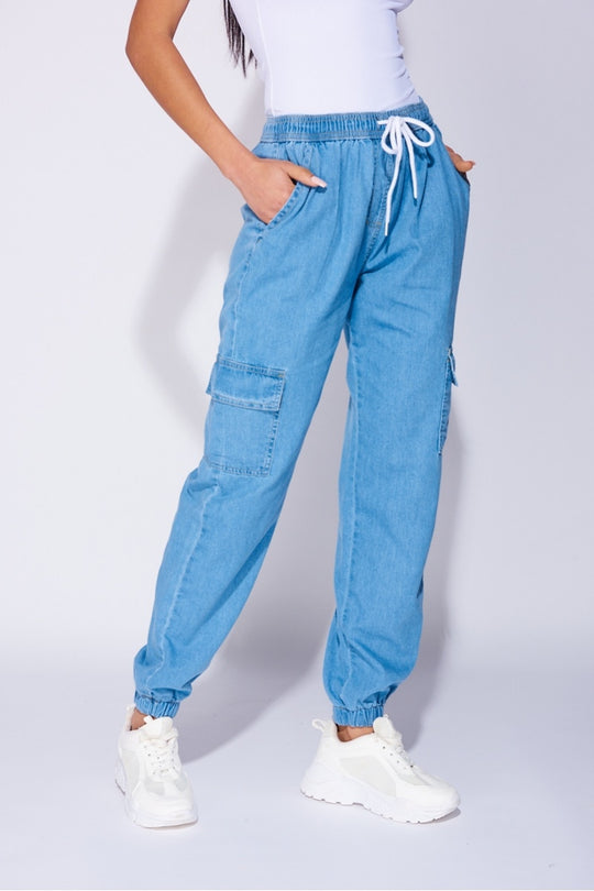 Model wears a loose fit light blue patch pocket cuffed denim joggers. Model wears a white bodysuit and white trainers.  Close up of the joggers sowing the model stands=ing slightly to the side. 