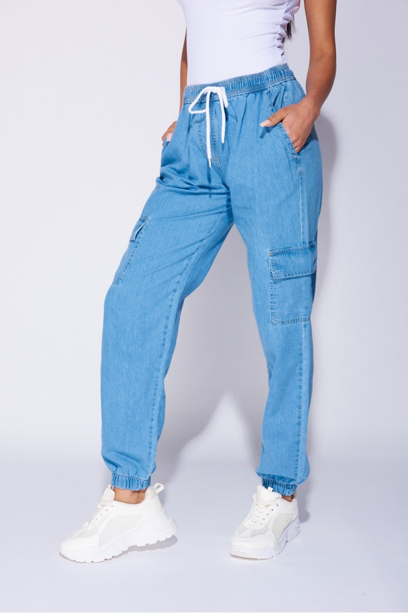 Model wears a loose fit light blue patch pocket cuffed denim joggers. Model wears a white bodysuit and white trainers.  Model stands to the side with their leg facing forward. 