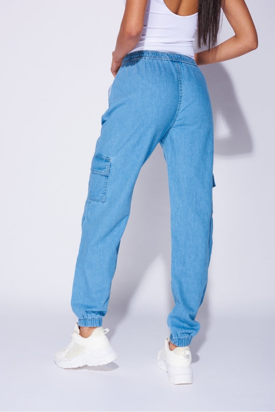 Model wears a loose fit light blue patch pocket cuffed denim joggers. Model wears a white bodysuit and white trainers. Closeup of the joggers with the model with her back to the camera, showing the back of the joggers.