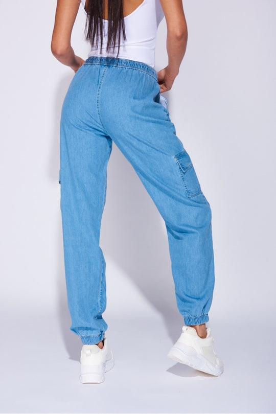 Model wears a loose fit light blue patch pocket cuffed denim joggers. Model wears a white bodysuit and white trainers.  Close up of the joggers show tghe back of the trousers. Model raises one leg. 