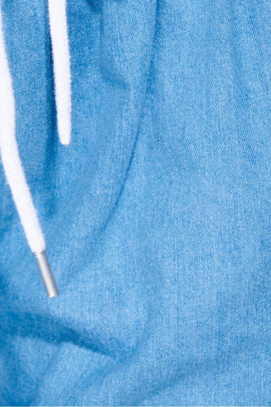 Model wears a loose fit light blue patch pocket cuffed denim joggers. Model wears a white bodysuit and white trainers. Photograph shows a closeup of the denim material and the white draw string detailing. 