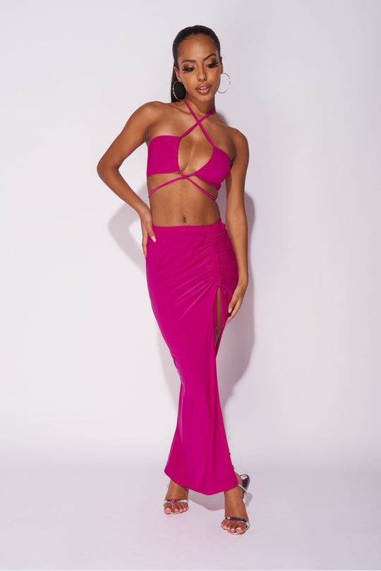 Model wears a magenta co-ord set. The top has a plunging criss cross crop top and the skirt has a thigh split and length adjusting string.   Model places one hand by her side and the other on her hip. 