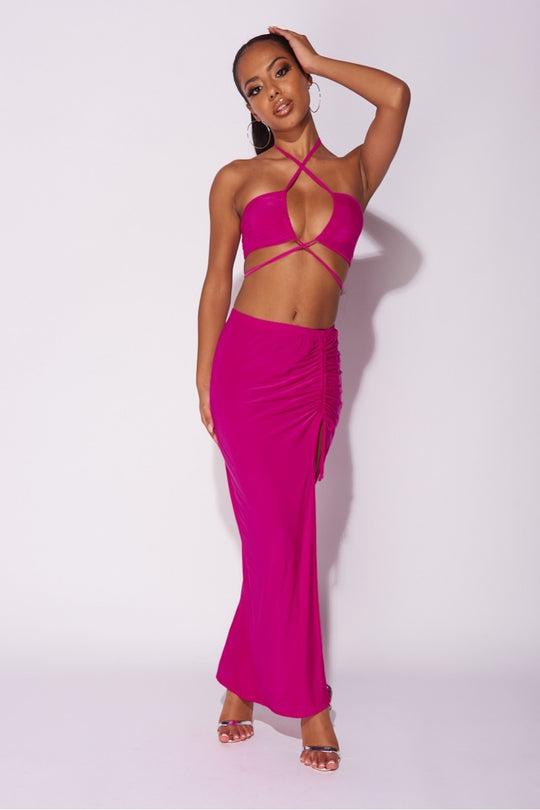 Model wears a magenta co-ord set. The top has a plunging criss cross crop top and the skirt has a thigh split and length adjusting string.  Model places one hand by her side, the other on her head and stands with her legs crossed. 