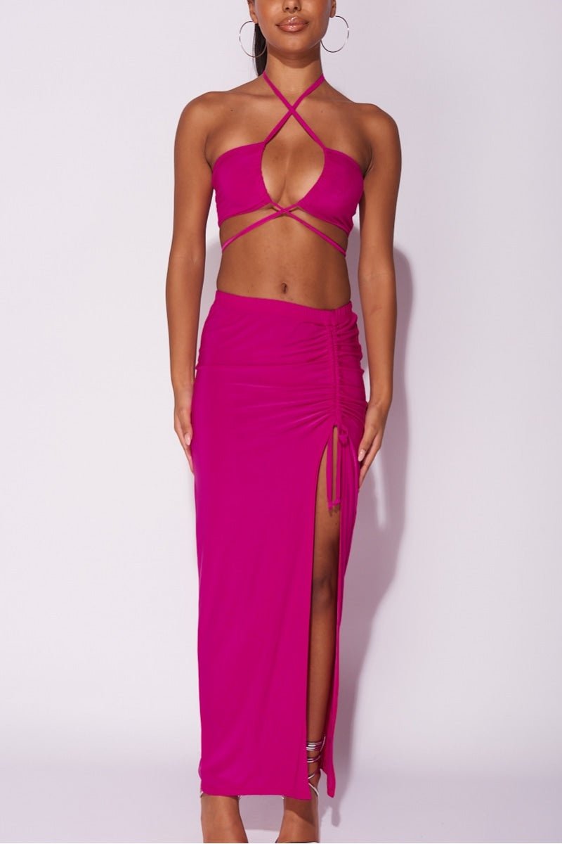 Model wears a magenta co-ord set. The top has a plunging criss cross crop top and the skirt has a thigh split and length adjusting string.  Model places both hands by her side. 