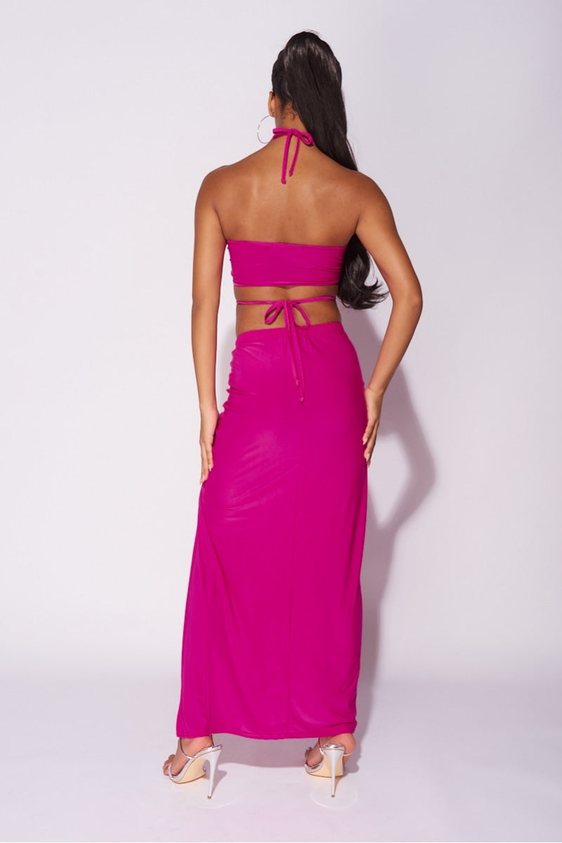 Model wears a magenta co-ord set. The top has a plunging criss cross crop top and the skirt has a thigh split and length adjusting string.  Model has her back to the camera, the back of the co-ord set is visible. 