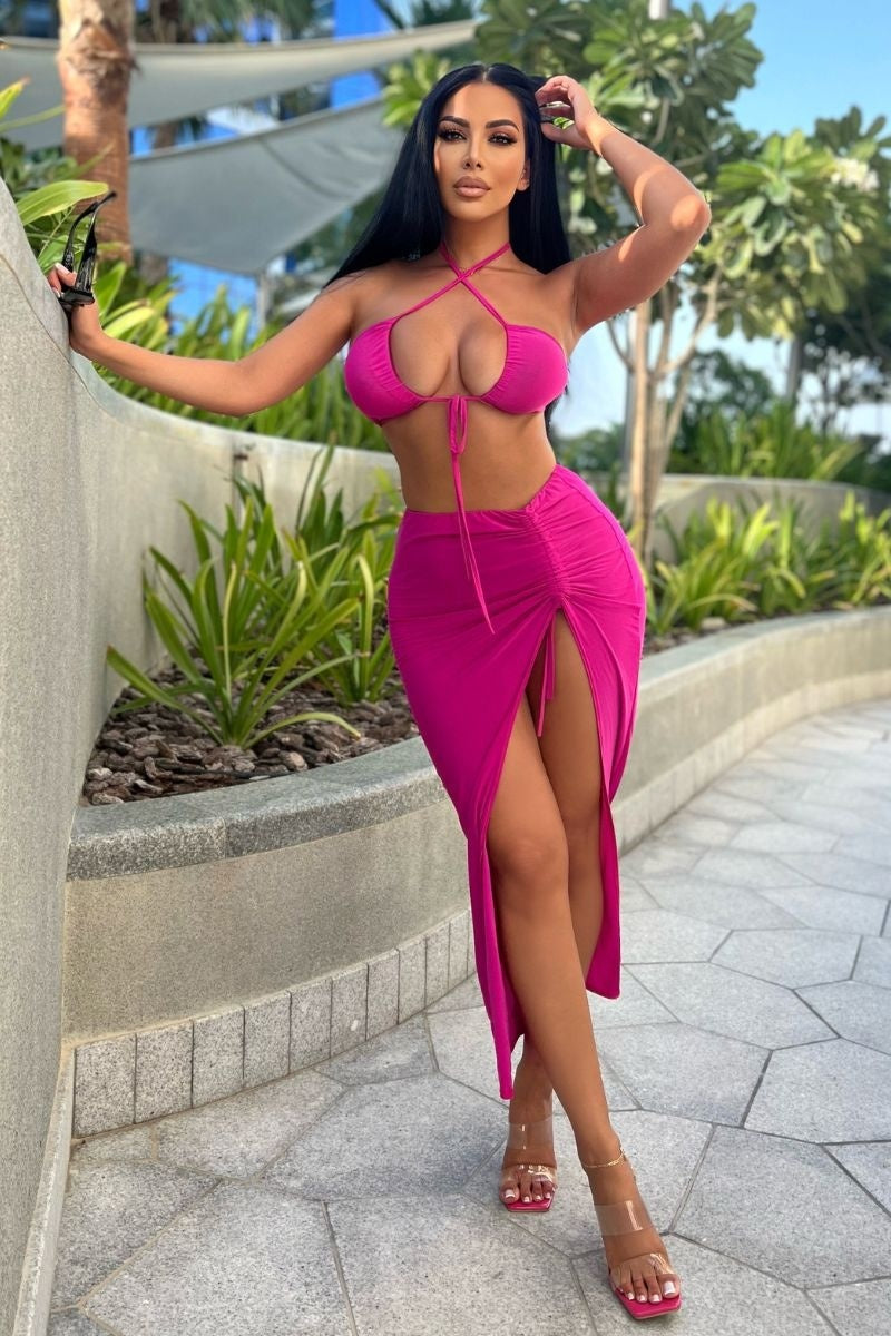 Model wears a magenta co-ord set. The top has a plunging criss cross crop top and the skirt has a thigh split and length adjusting string.  Model is outside and places her hand on a wall and the other by her head.