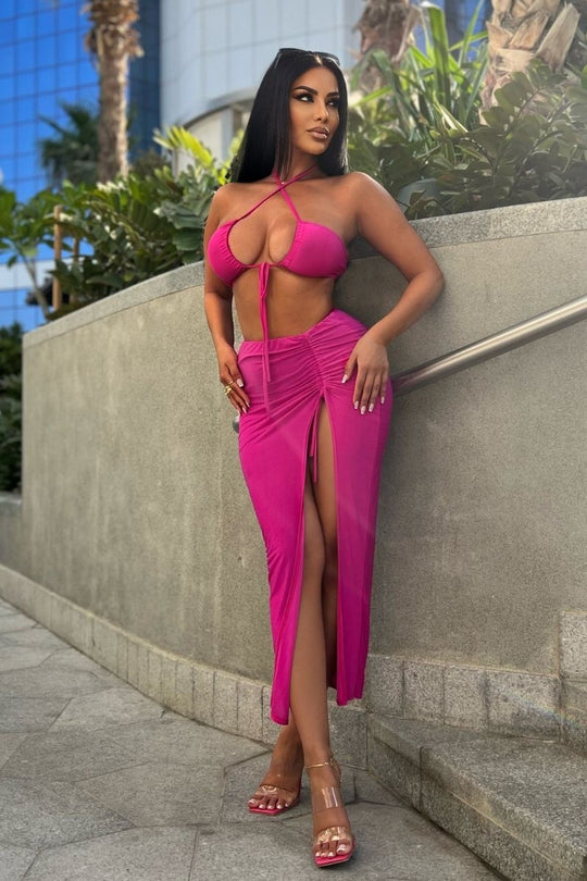 Model wears a magenta co-ord set. The top has a plunging criss cross crop top and the skirt has a thigh split and length adjusting string.  Model is outside and leans against a wall.