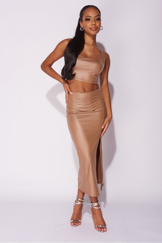 Model wears faux leather co-ordinated set in mocha. The crop top has a deep crew neck and the midi skirt has a thigh high split.  Model has one arm on her thigh and one on her hip and stares into the camera. 