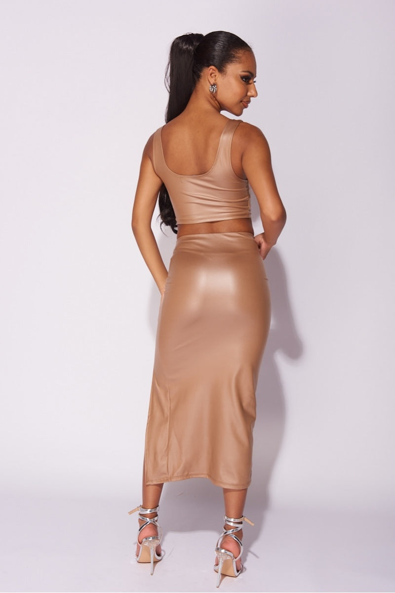Model wears faux leather co-ordinated set in mocha. The crop top has a deep crew neck and the midi skirt has a thigh high split.  Model has her back to the camera  and looks to the side.