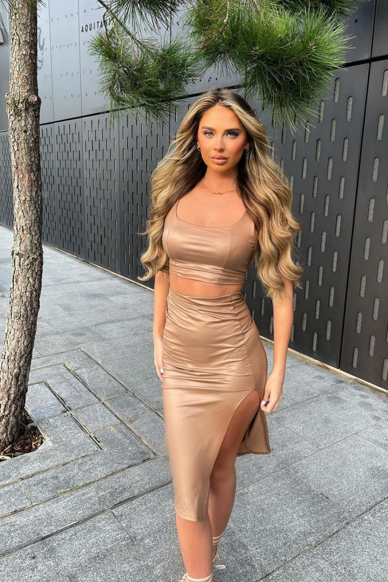 Model wears faux leather co-ordinated set in mocha. The crop top has a deep crew neck and the midi skirt has a thigh high split.  Model stares directly into the camers aith her hands by her sides. and one leg crossed in front of the over. 