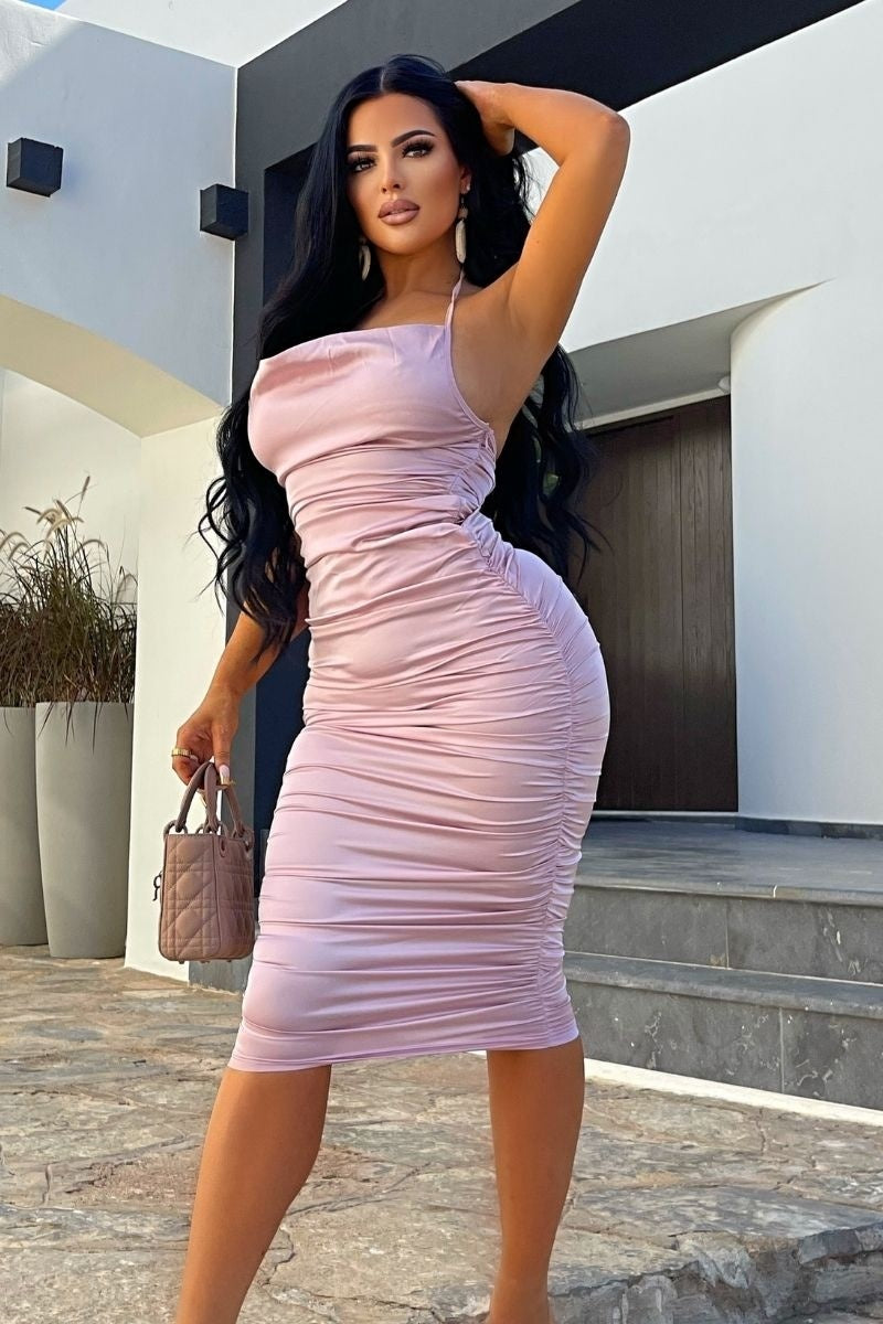 Model wears pink satin midi dress, with a cowl neck and thinned straped halter neck detailing. The dress has ruched detailing at the front and rear of the dress. Model holds a bag in one hand and her hand above her head with the other. 