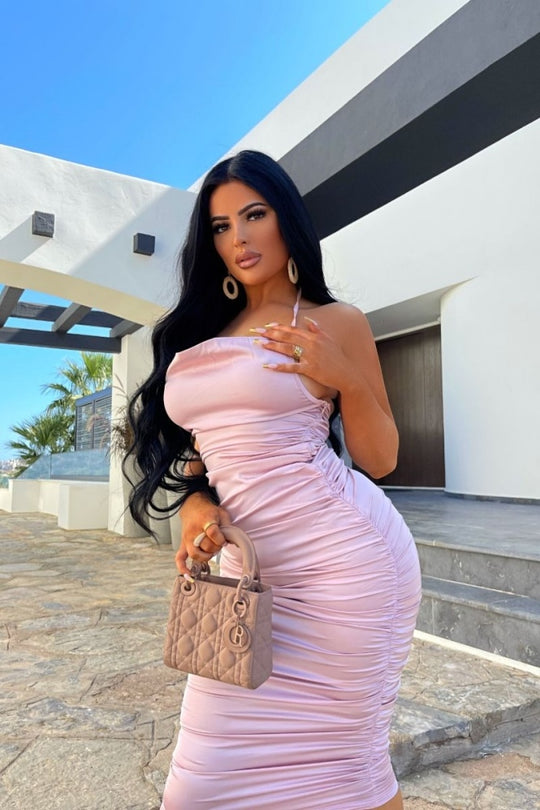 Model wears pink satin midi dress with cowl neck design. The dress has ruched sides and a concealed zip fastening. Model holds a handbag in one hand and touches the strap of her dress with the other.